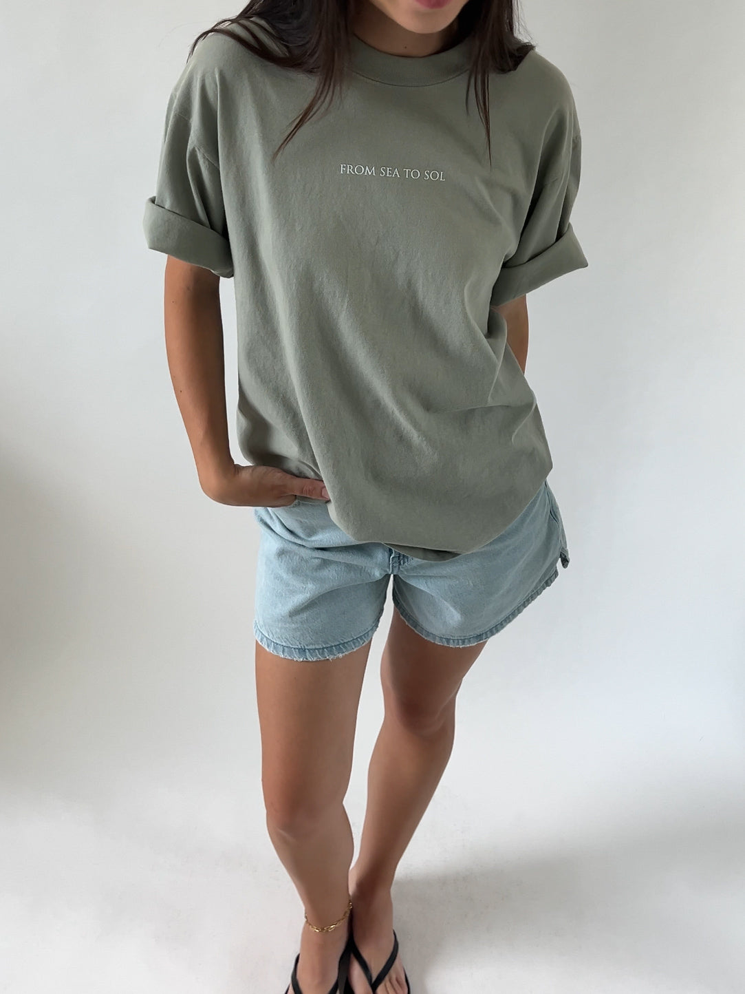 A person wearing a green t-shirt with the text 'FROM SEA TO SOL' on the front and blue denim shorts, standing against a white background.