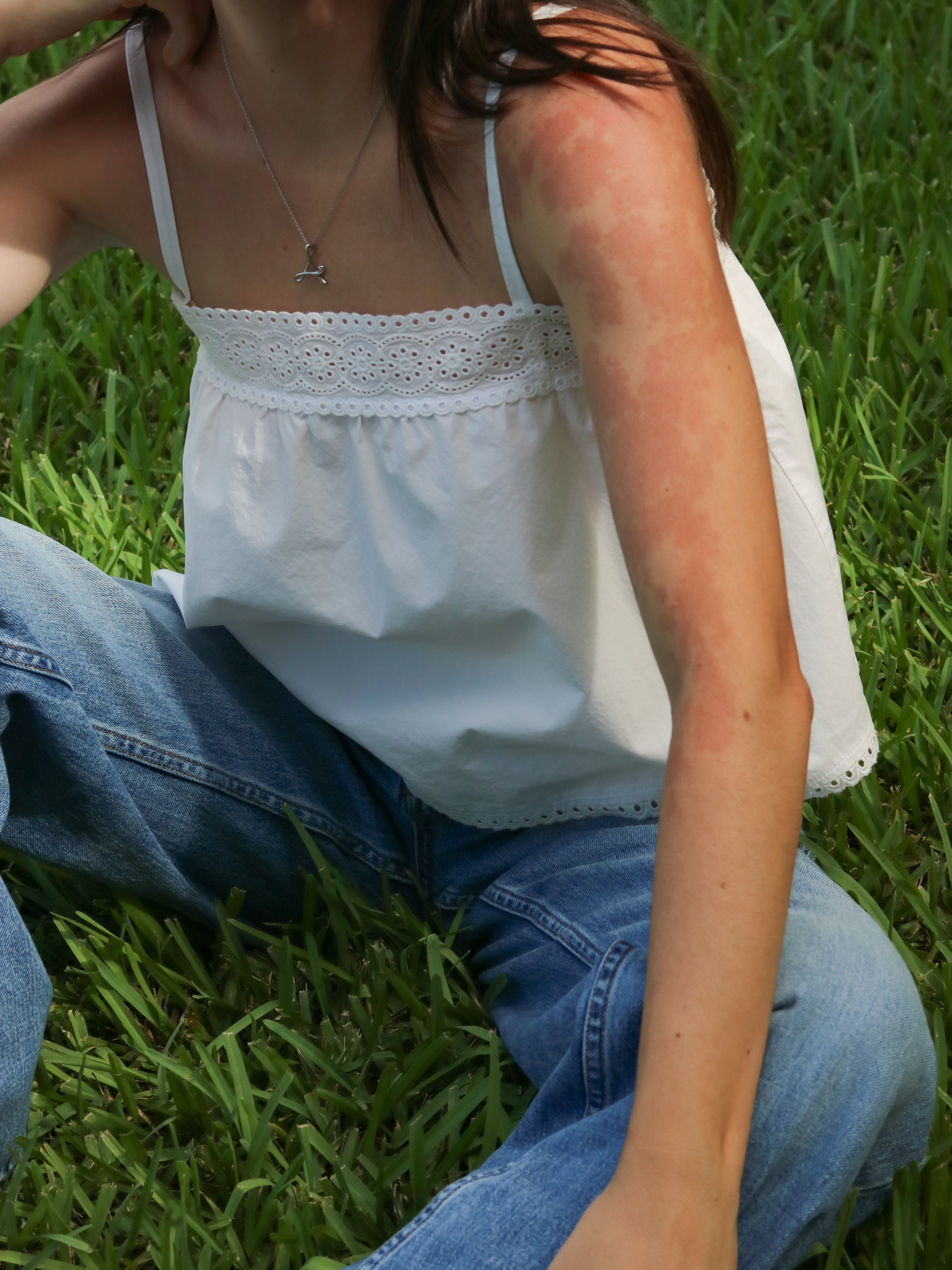 A woman sitting on green grass wearing a white top with thin straps and eyelet lace trim, paired with blue jeans.