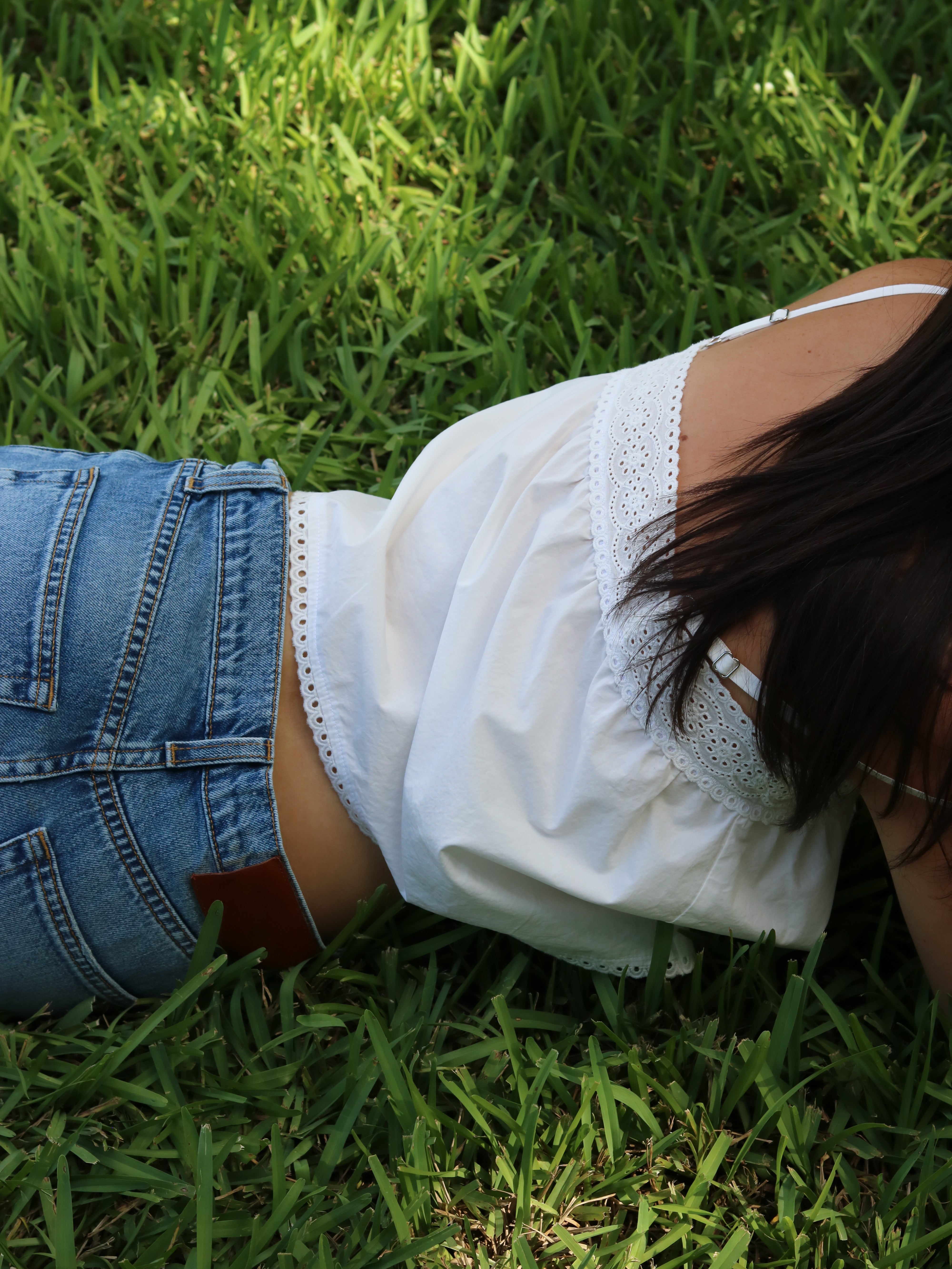 A person lying on green grass wearing a white eyelet-embroidered camisole top and blue denim shorts.