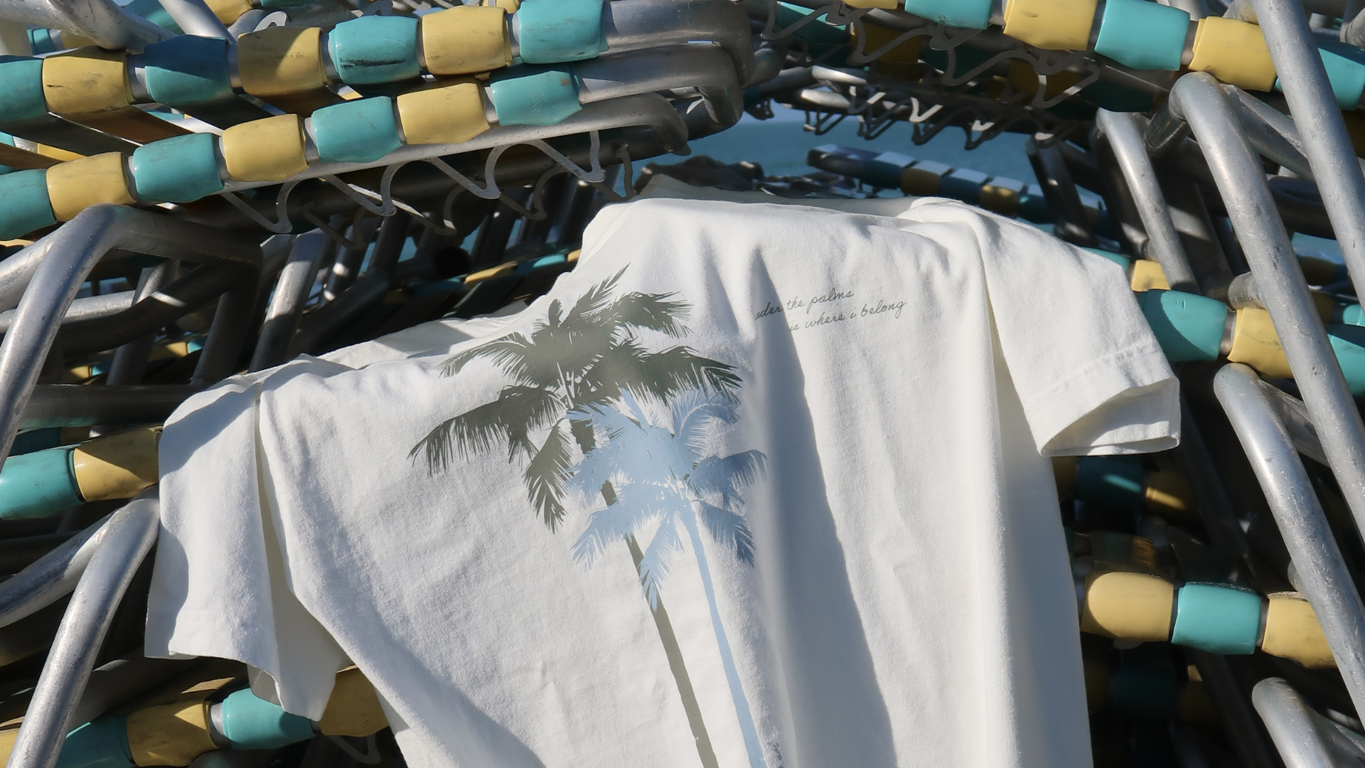 White t-shirt with palm tree design on a rack of folded beach chairs.