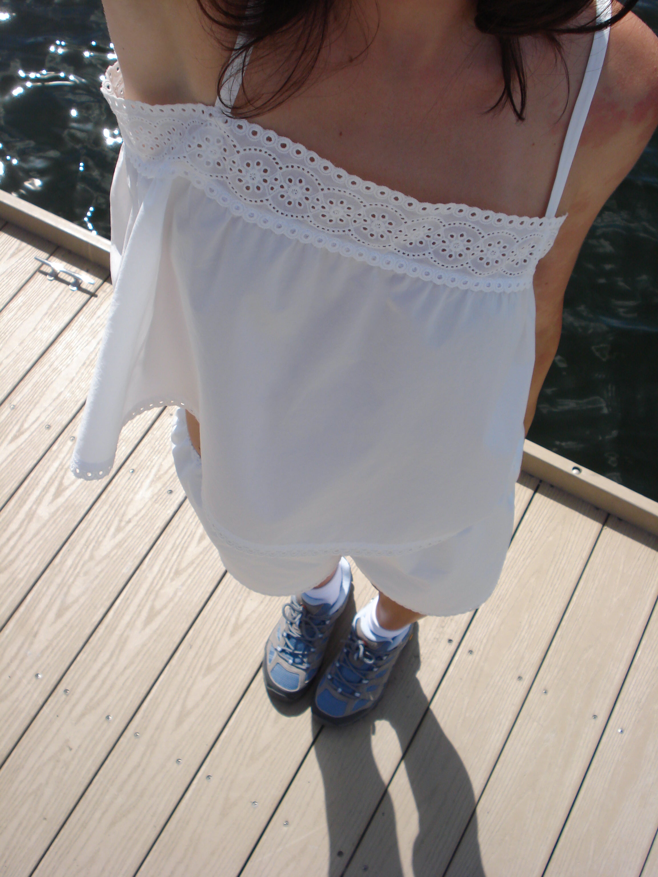 A person standing on a wooden dock wearing a white spaghetti-strap top with a crochet eyelet neckline and white shorts, paired with blue sneakers.