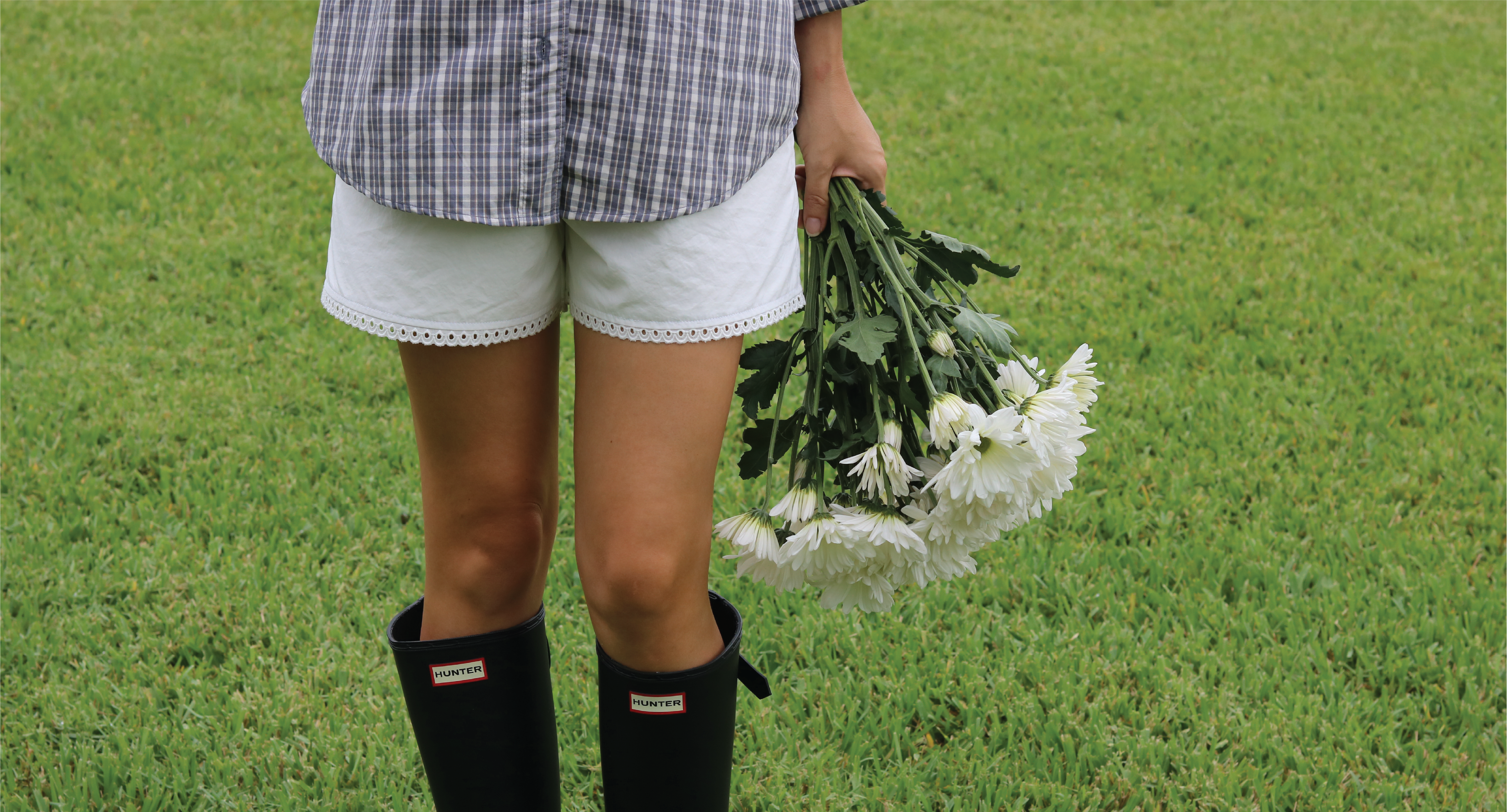 A person standing on a green grassy lawn wearing a blue and white plaid shirt, white shorts with lace trim, black Hunter rain boots, and holding a bouquet of white daisies.