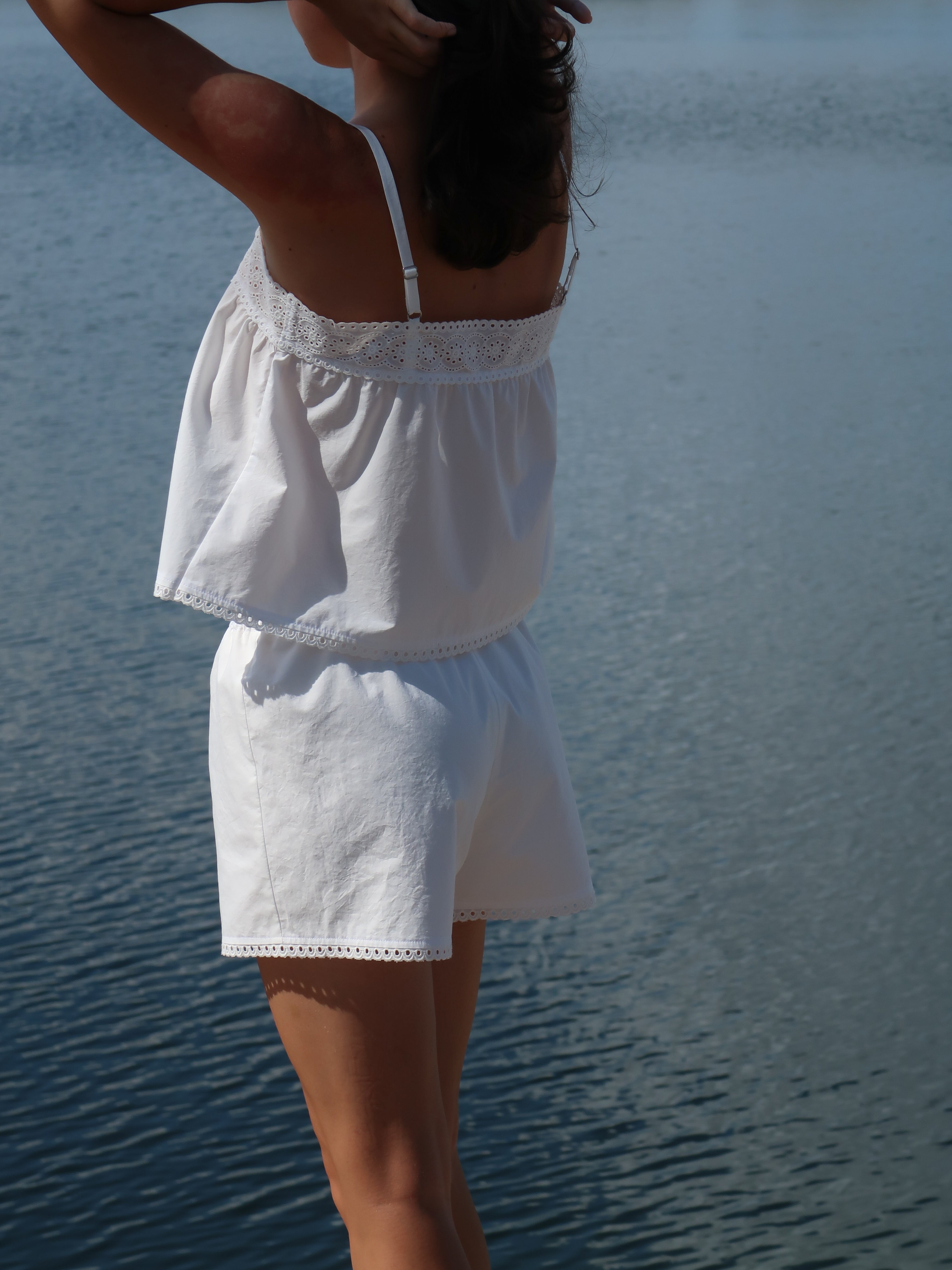 A woman wearing a white two-piece set with lace trim, consisting of a camisole top and matching shorts, standing outdoors by a body of water.